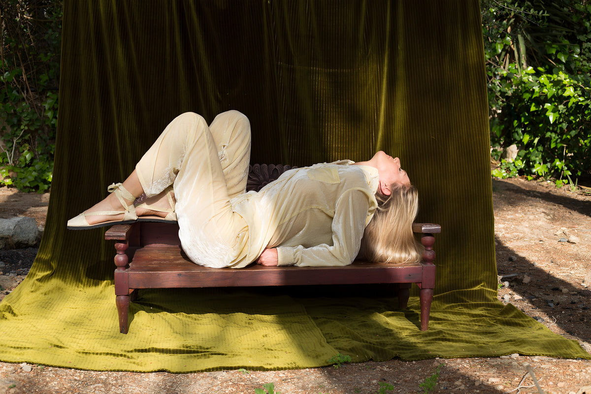 Model in a cream La Fleury velvet set lying on a wooden daybed in front of an olive velvet curtain