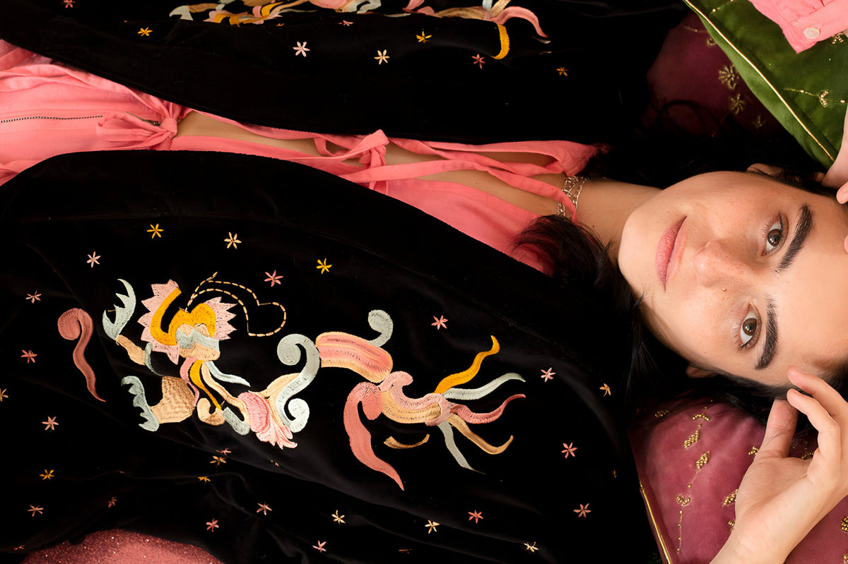 Model lying on embroidered velvet cushions wearing a black velvet kimono hand-embroidered with dragons and stars