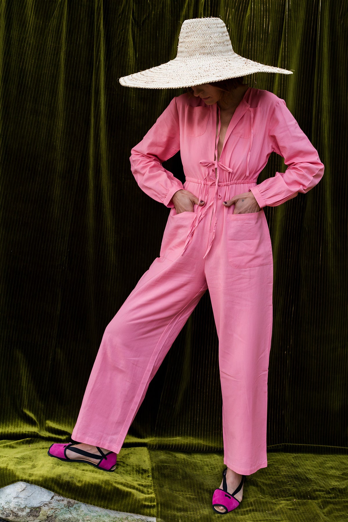 Model in a pink jumpsuit and a wide straw hat in front of an olive velvet curtain