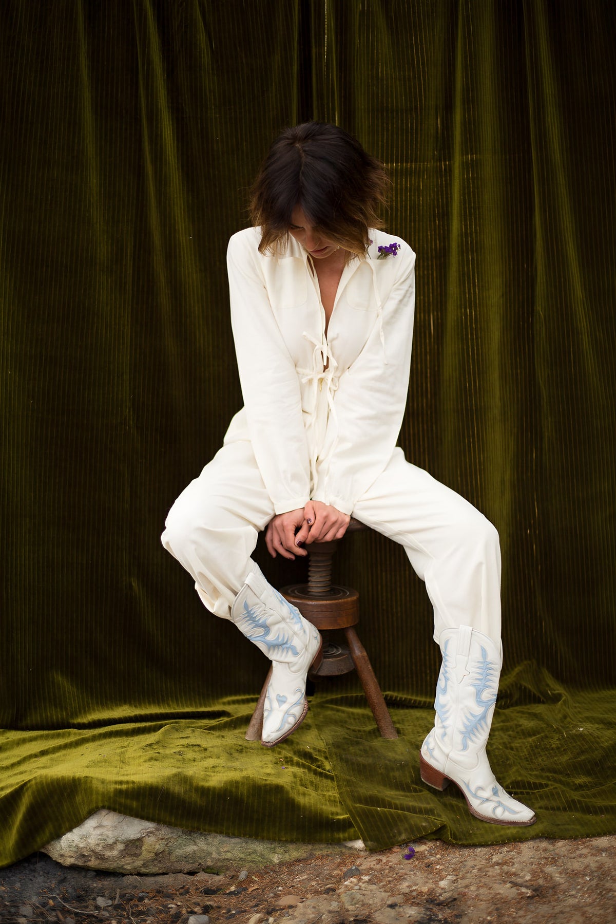 Model in a white suit and embroidered cowboy boots sitting on a wooden stool in the garden